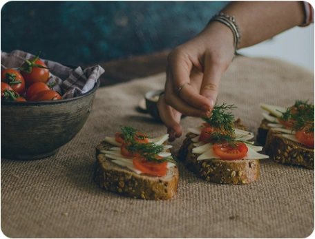 Hand placing herbs on rye bread with tomatoes on a wooden table